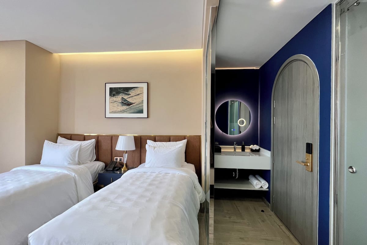 A hotel room with two beds, a bedside table with a lamp, a painting on the wall, a bathroom with a sink and mirror, and a wardrobe.