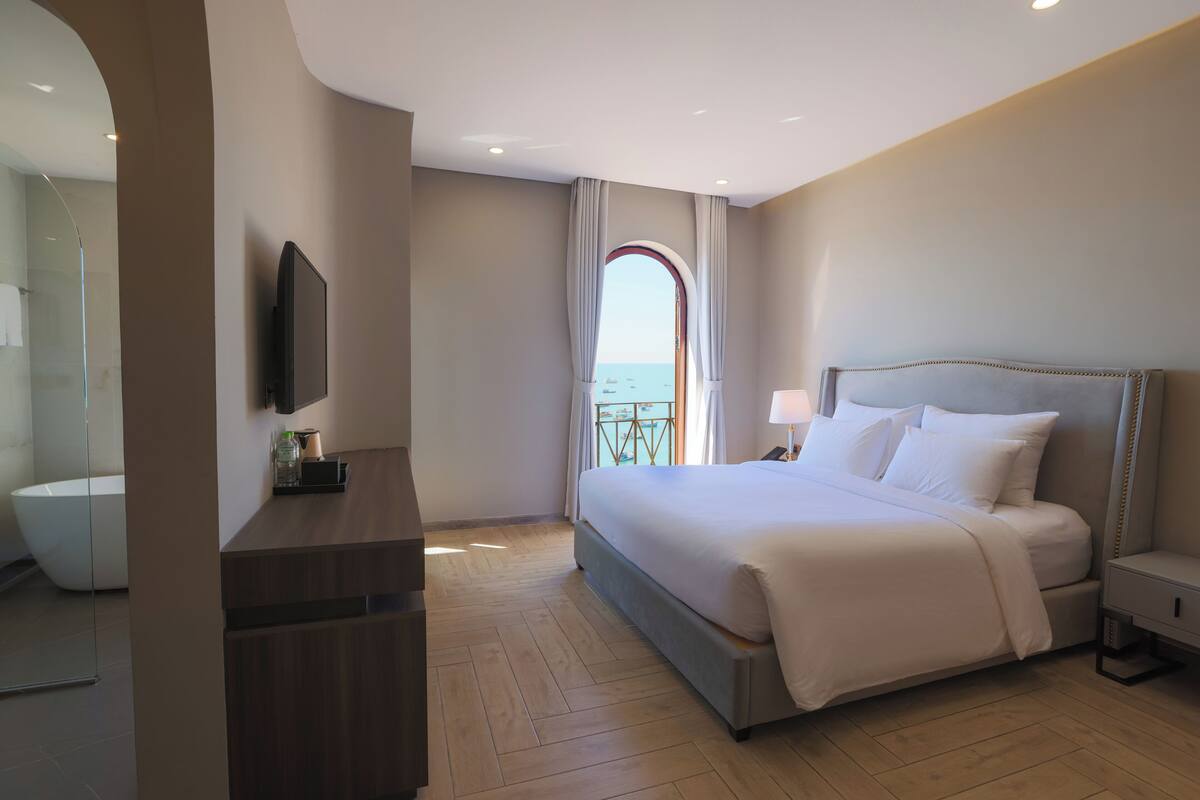 A hotel room with a large bed, a TV mounted on the wall, a bedside table, and a view of the sea through a window.