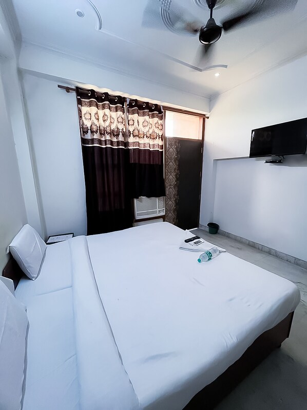 Habitación de hotel con una cama individual, ventilador de techo, televisión montada en la pared y una ventana con cortinas estampadas.