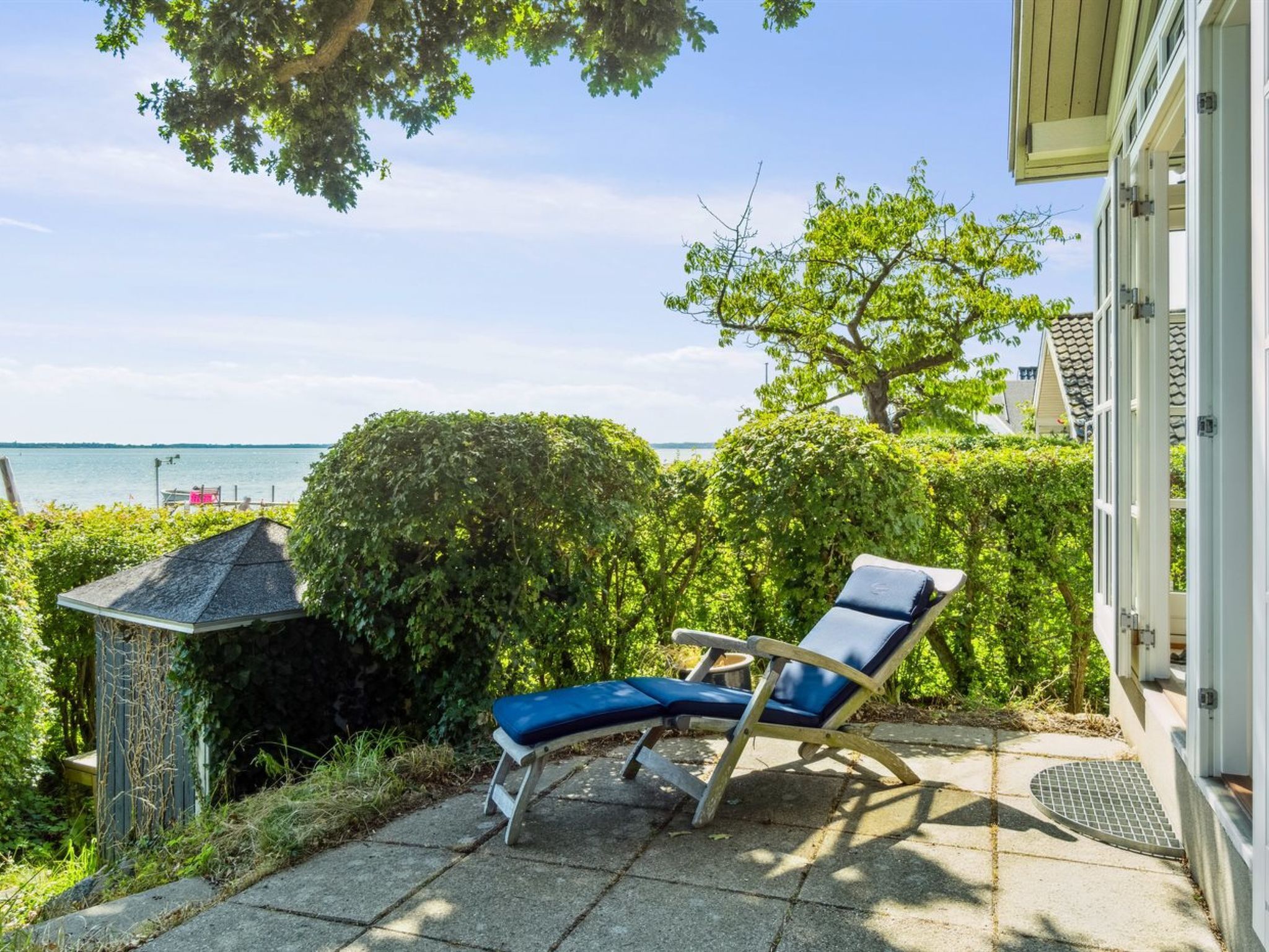 Een zonnig terras met een lounge stoel, een kleine schuur en uitzicht op het water.