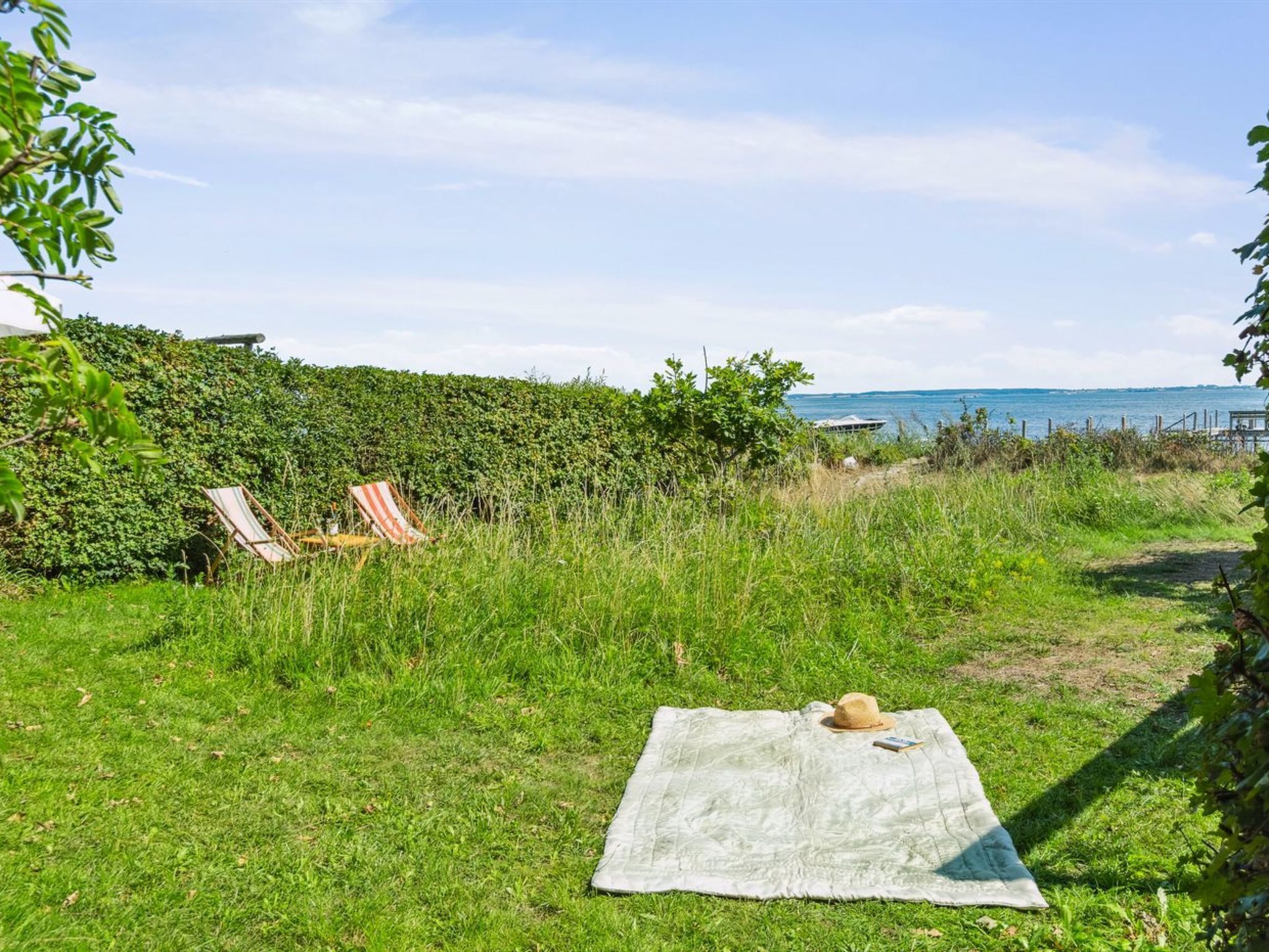 A grassy outdoor area with a picnic setup including a blanket and a hat, a chair, and a hedge in the background.