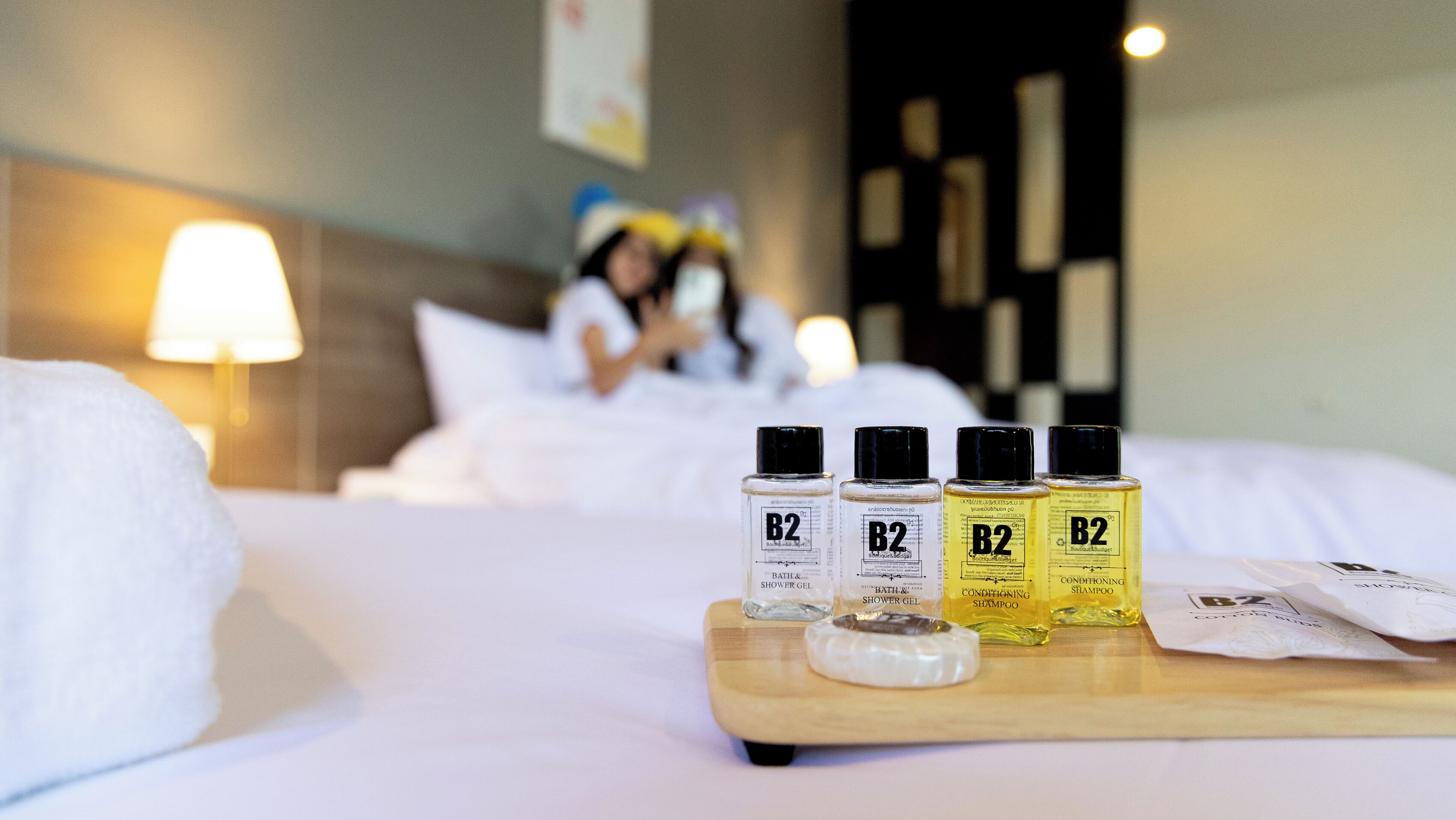 Et hotelværelse med en pænt redt seng, en bakke med fire flasker B2 bade- og brusegel, samt en flaske B2 conditioner shampoo.