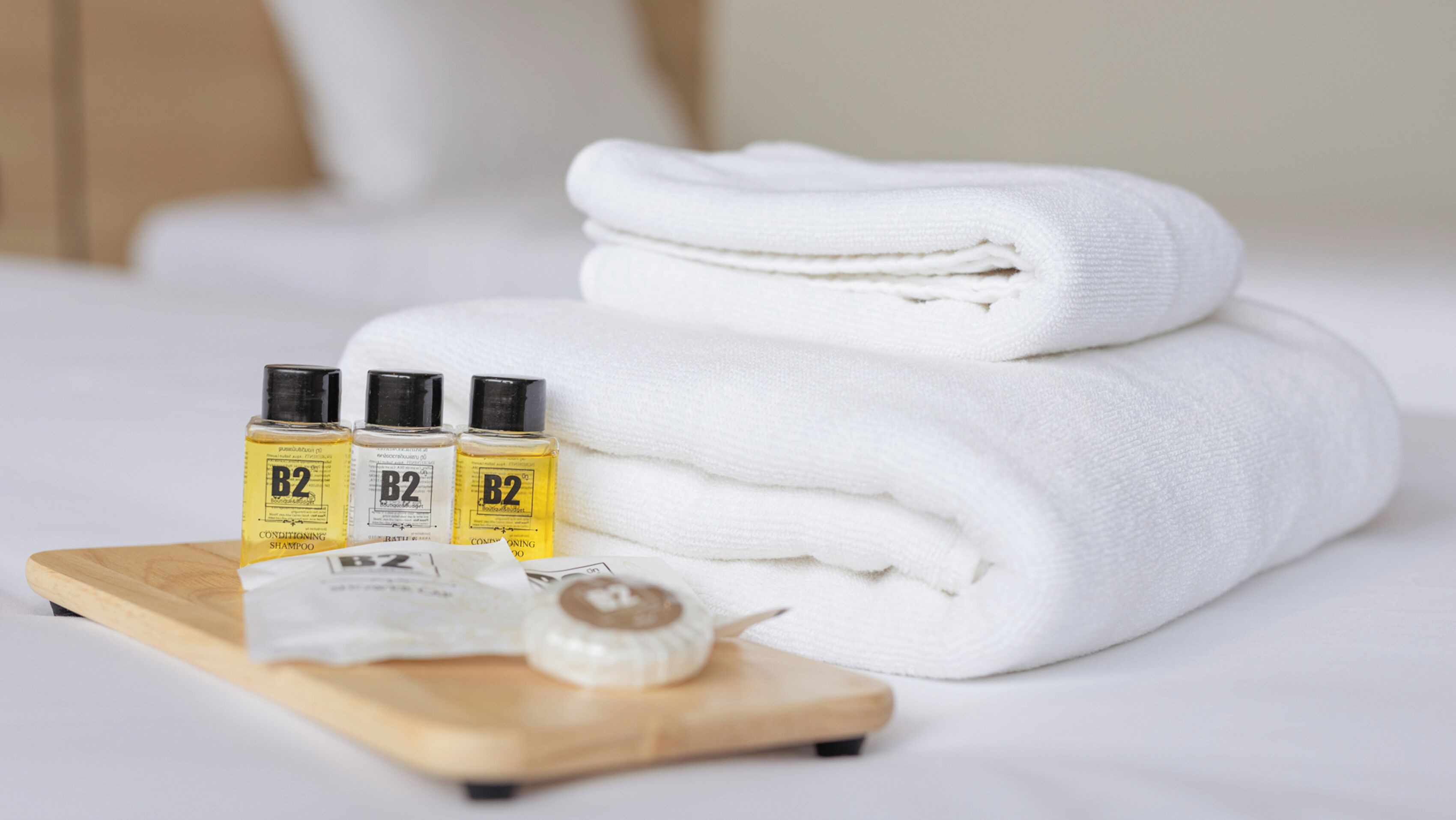 A neatly folded white towel stacked on a bed, three bottles of B2 conditioning shampoo, and a wooden tray with a small item on it.