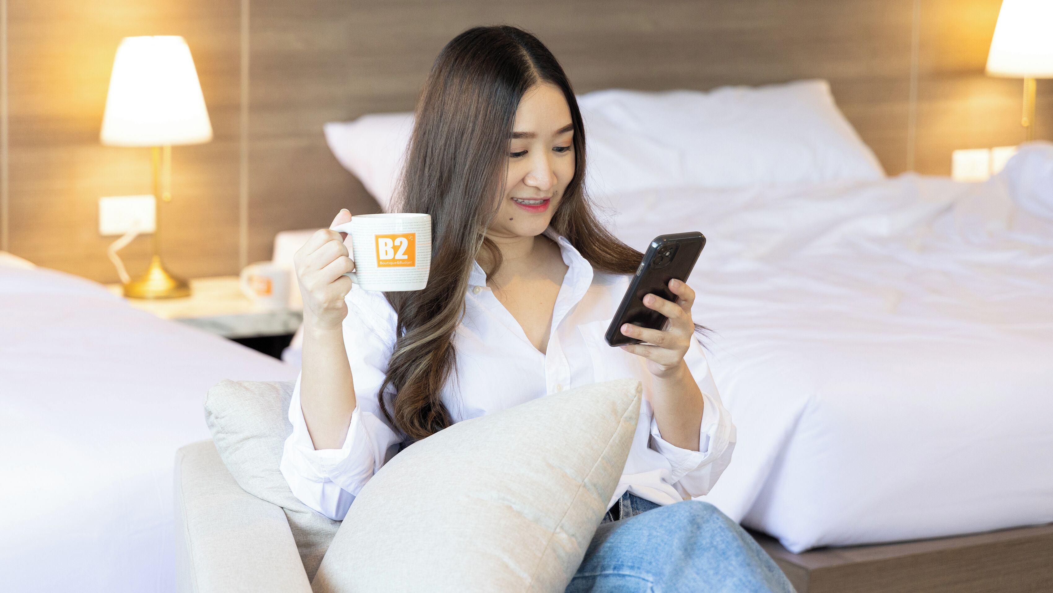 A person sitting on a bed in a hotel room, holding a smartphone and a mug.