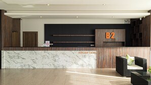 Un espace d’accueil doté d’un comptoir en marbre, d’un panneau indiquant « B2 » et d’un coin salon agrémenté d’une plante.