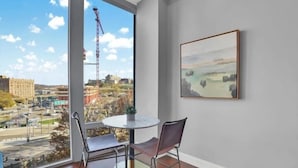A room with a large window offering a view of a construction site and cityscape. There is a round table with two chairs and a framed painting on the wall.