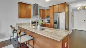 A modern kitchen with wooden cabinets, a marble countertop, stainless steel appliances, and a bar area with stools.