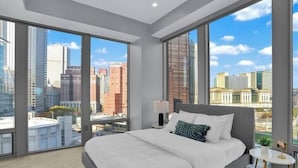 A modern hotel room with a large bed, a nightstand, and a view of the cityscape through floor-to-ceiling windows.