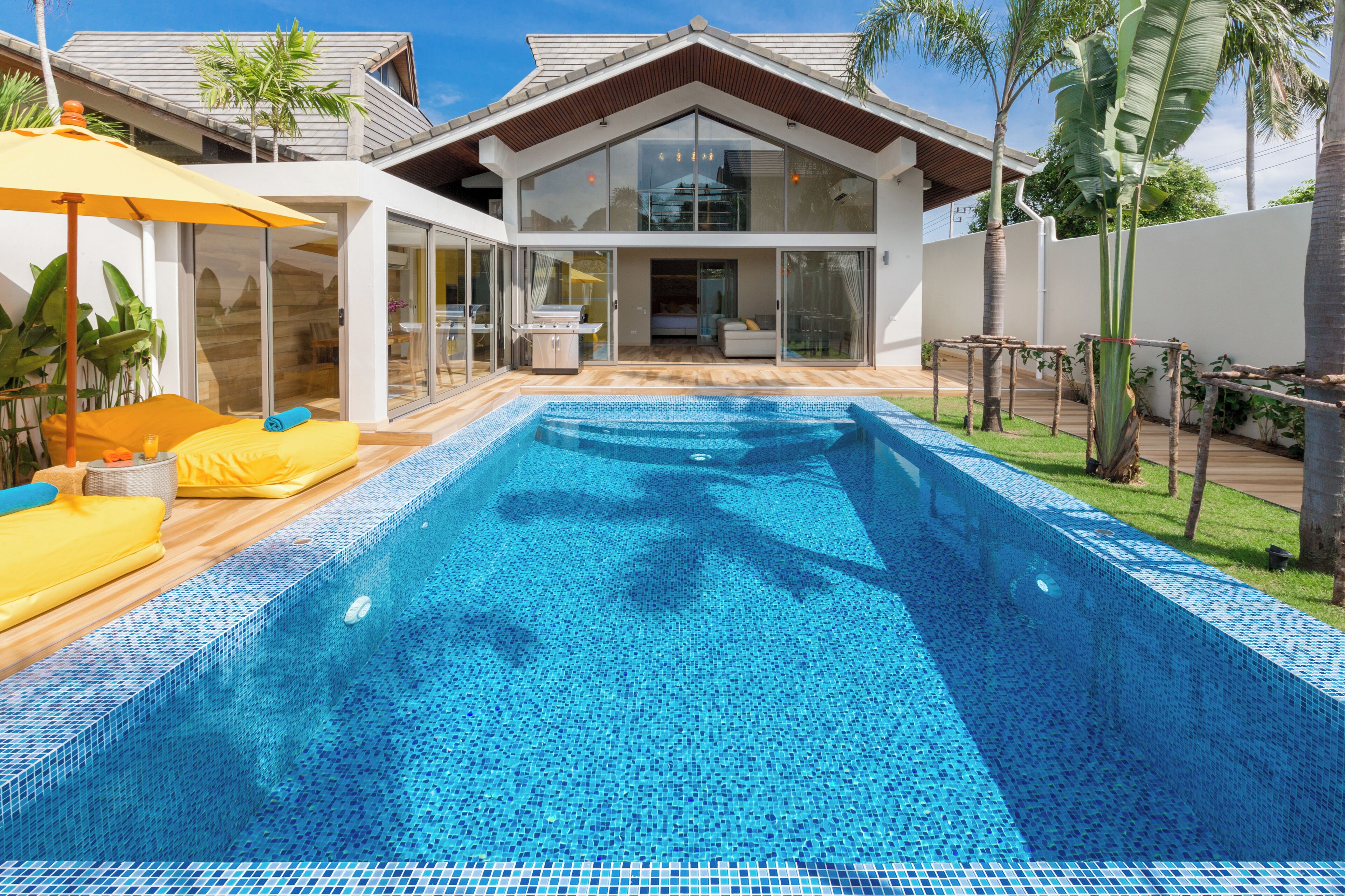Une maison moderne à deux étages, dotée d’une grande façade vitrée, d’une piscine et d’un patio agrémenté de coussins jaunes.