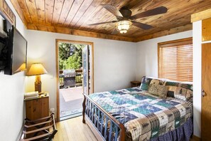 Un dormitorio con techo de madera, una cama con colcha, una mesita de noche, una lámpara y una puerta que da a un espacio exterior.