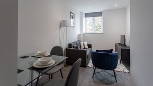 En moderne stue med et glasbord, en grå sofa, en blå lænestol og et stort vindue der giver naturligt lys.