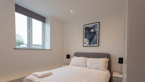 Et soveværelse med en stor seng, et vindue, et natbord, en lampe og et indrammet abstrakt billede på væggen.