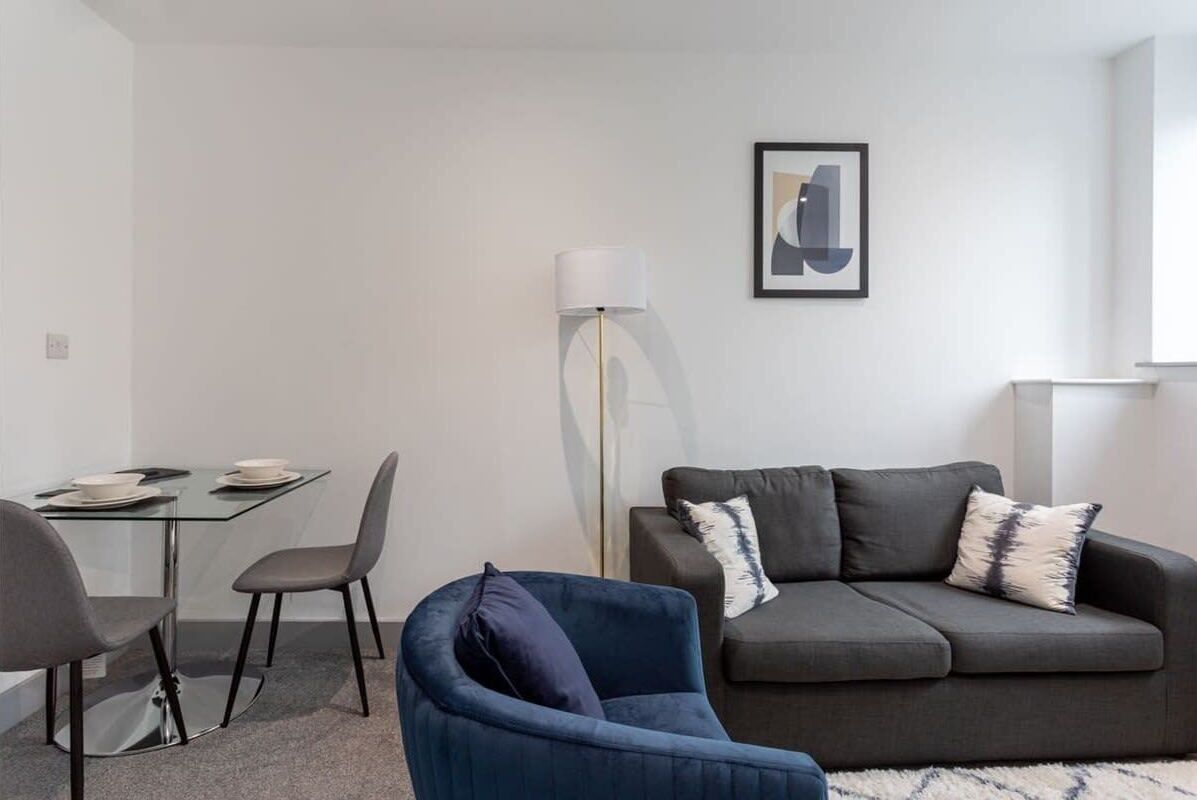Una sala de estar moderna con una mesa de comedor de vidrio, dos sillas, un sillón azul, un sofá gris con cojines estampados y una lámpara de pie.