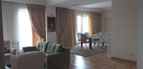 Una sala de estar con sofá, sillas, una mesa de comedor con sillas y un gran ventanal con cortinas.