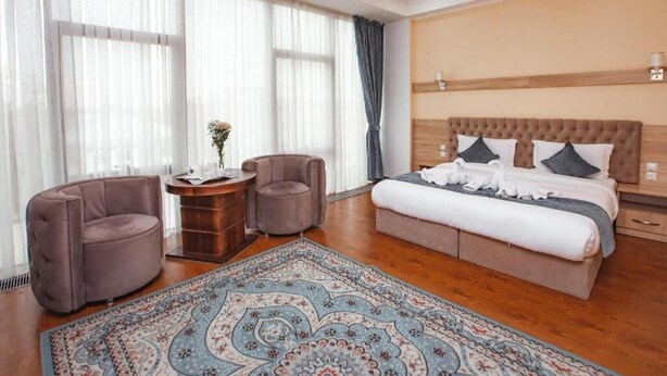 A hotel room with a large bed, two armchairs, a wooden side table with a vase of flowers, and a patterned rug. - Park Hotel al Bustan (Shymkent)