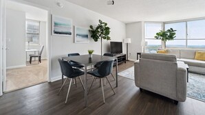 A modern living room with a dining area, a grey sofa, a flat-screen TV, and large windows offering a view. - 2BR Executive City Suite With Gym & Pool (Arlington)