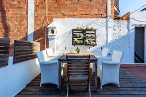 Une terrasse avec une table en bois et des chaises en osier blanc, un mur de briques et un bâtiment blanc doté d’une fenêtre et d’une porte.