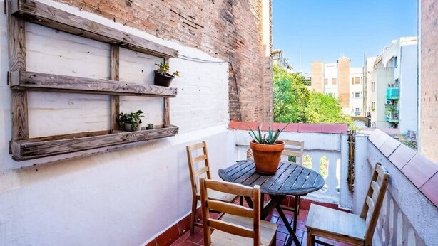 A small balcony with a wooden table and chairs, potted plants, and a white wall with exposed brick. - Private terrace Attico 3BD in Gran de Gracia (Barcelona)
