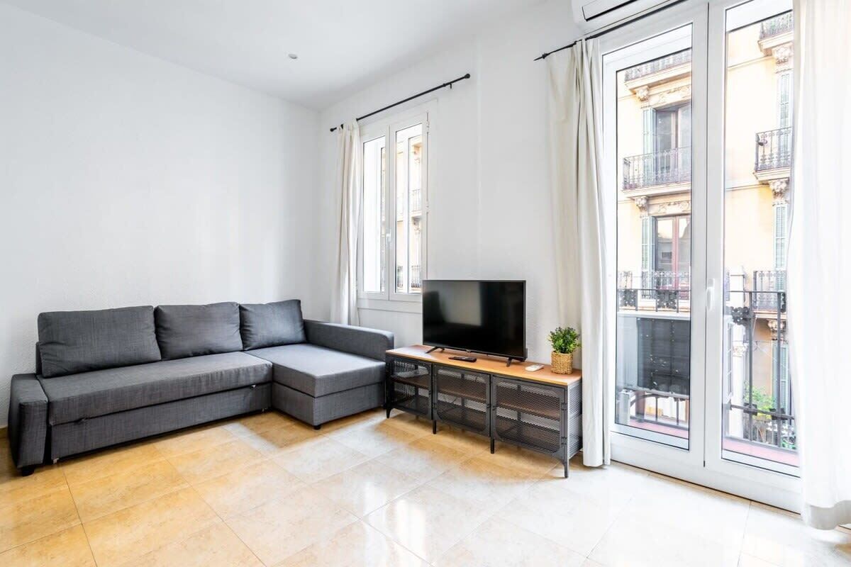 Un soggiorno moderno con un divano angolare grigio, un televisore a schermo piatto su un supporto in legno e un'ampia finestra con vista sulla città.