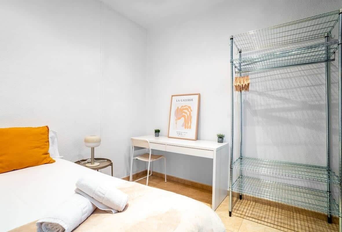 Une chambre d’hôtel comprenant un lit, un bureau, une chaise, une petite table avec une lampe, un tableau encadré au mur et une étagère en métal.