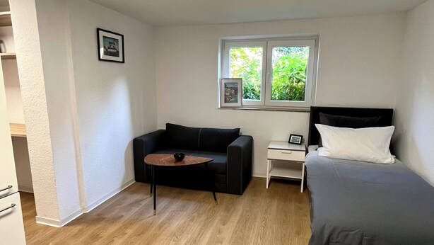 A compact room with a bed, a sofa, a small table, and a window with a view of greenery. - 05 Central & quiet with SmartTV (Fellbach)