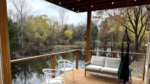 A wooden deck with a white sofa and two white chairs, overlooking a body of water and trees.