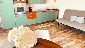 Una cocina compacta con horno empotrado, cafetera y hervidor en la encimera. En primer plano, una mesa de comedor con un florero. Un sofá con cojines está apoyado contra la pared, y se observa un suelo de madera.