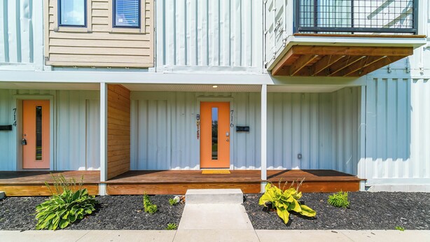 Un bâtiment moderne à deux étages doté d’une terrasse en bois, d’un petit jardin agrémenté de plantes et d’un chemin en béton menant à l’entrée.