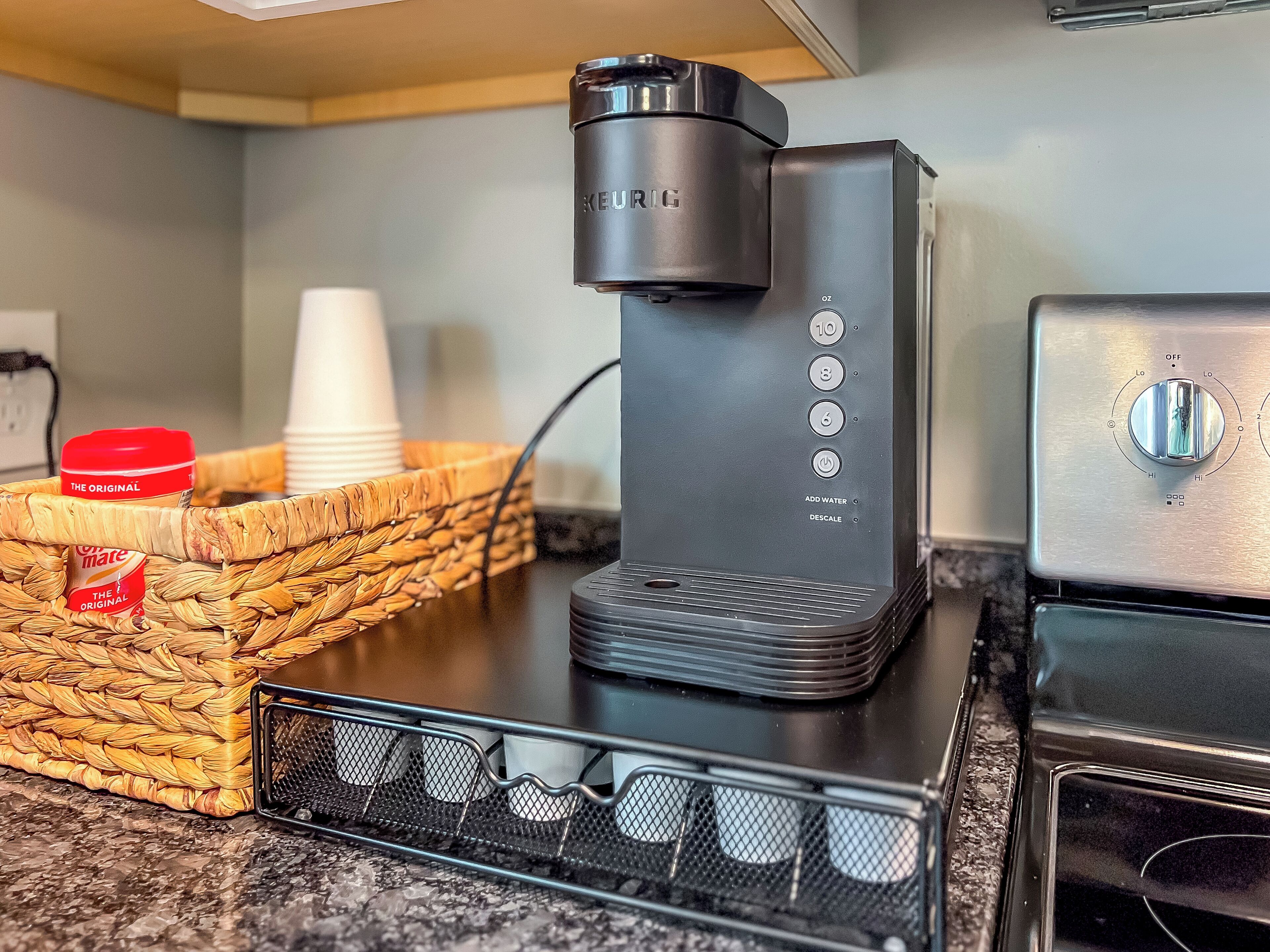 A Keurig coffee maker on a countertop with a basket of disposable cups and a jar of creamer.