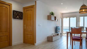 Amplia habitación con suelo de madera, zona de comedor con mesa y sillas, televisor sobre un mueble y un amplio ventanal con vistas al mar.