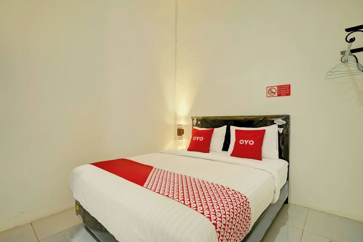 A neatly made bed with a white and red bedspread, two red pillows with the word "OYO" on them, and a small sign on the wall indicating no smoking.