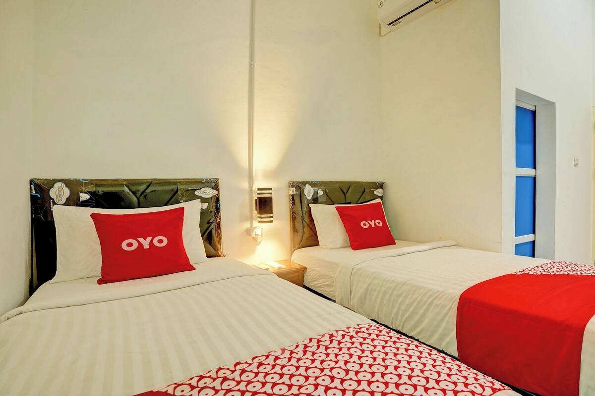 A hotel room with two beds, each with a red pillow labeled "OYO," a bedside table, and a wall-mounted light.