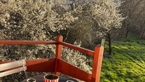 Ein Holzhocker mit einem runden Tisch, zwei gestreifte Tassen und Blick auf einen Garten mit blühenden Bäumen.