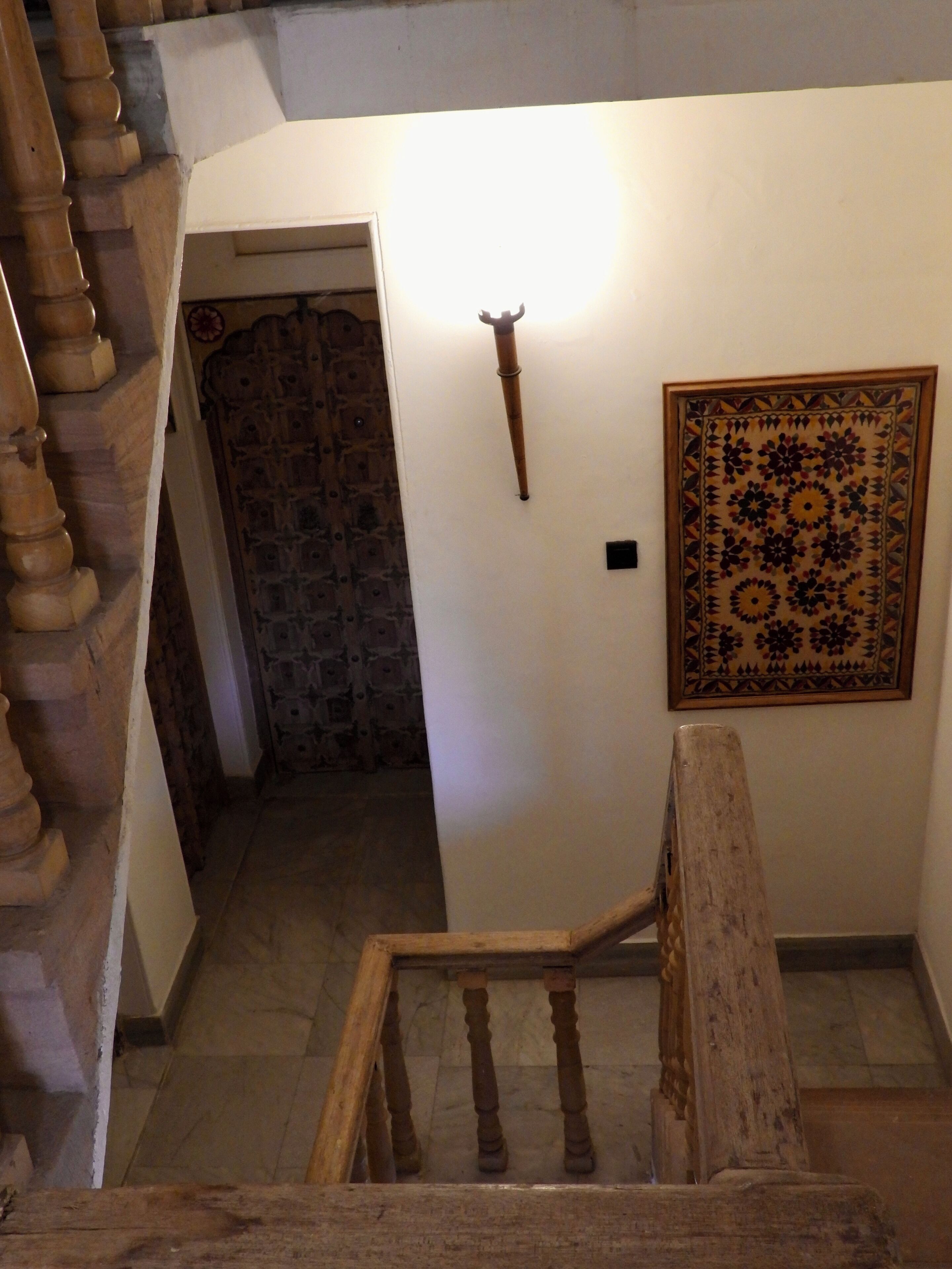 A staircase with wooden balusters leading to a room with a patterned wall hanging and a door with a stone wall behind it.