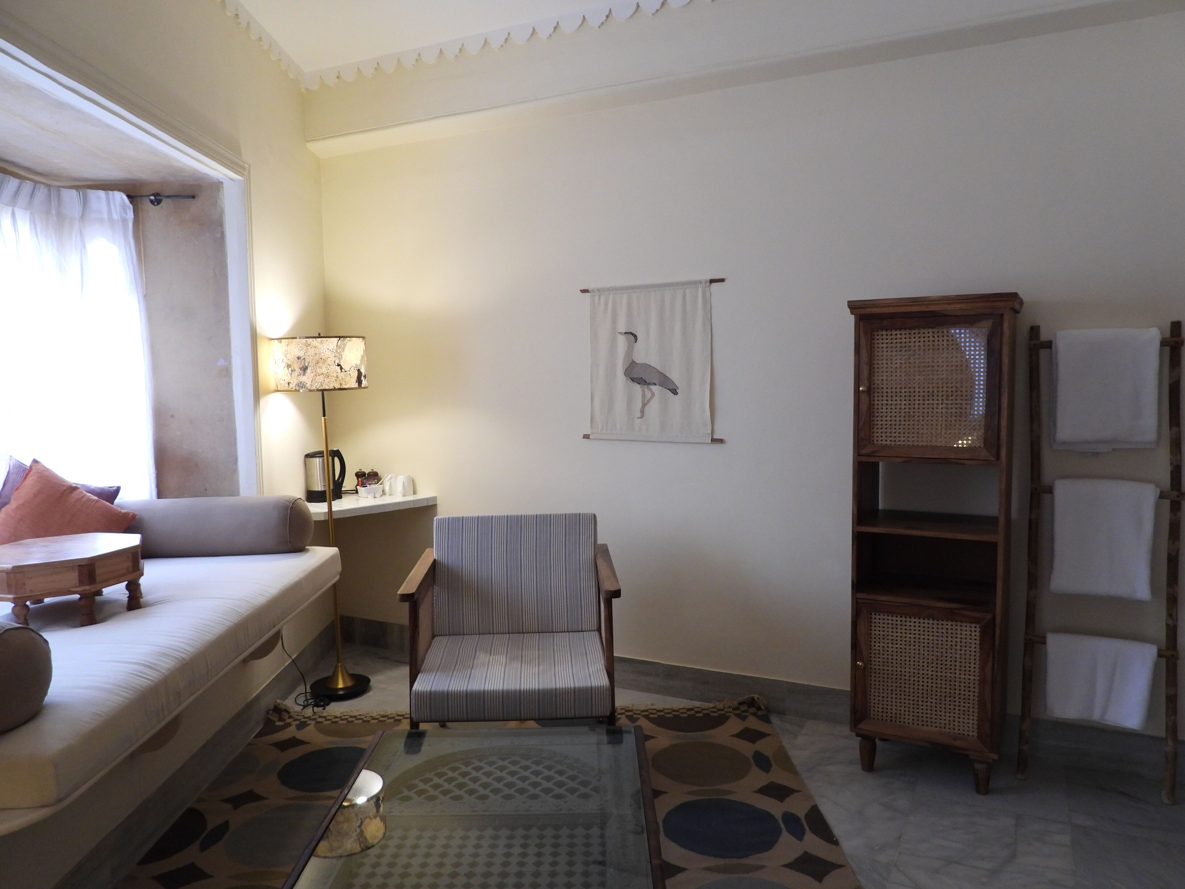 A room with a sofa, armchair, and a small table with a lamp. There is a wall-mounted shelf with towels and a framed picture of a bird.