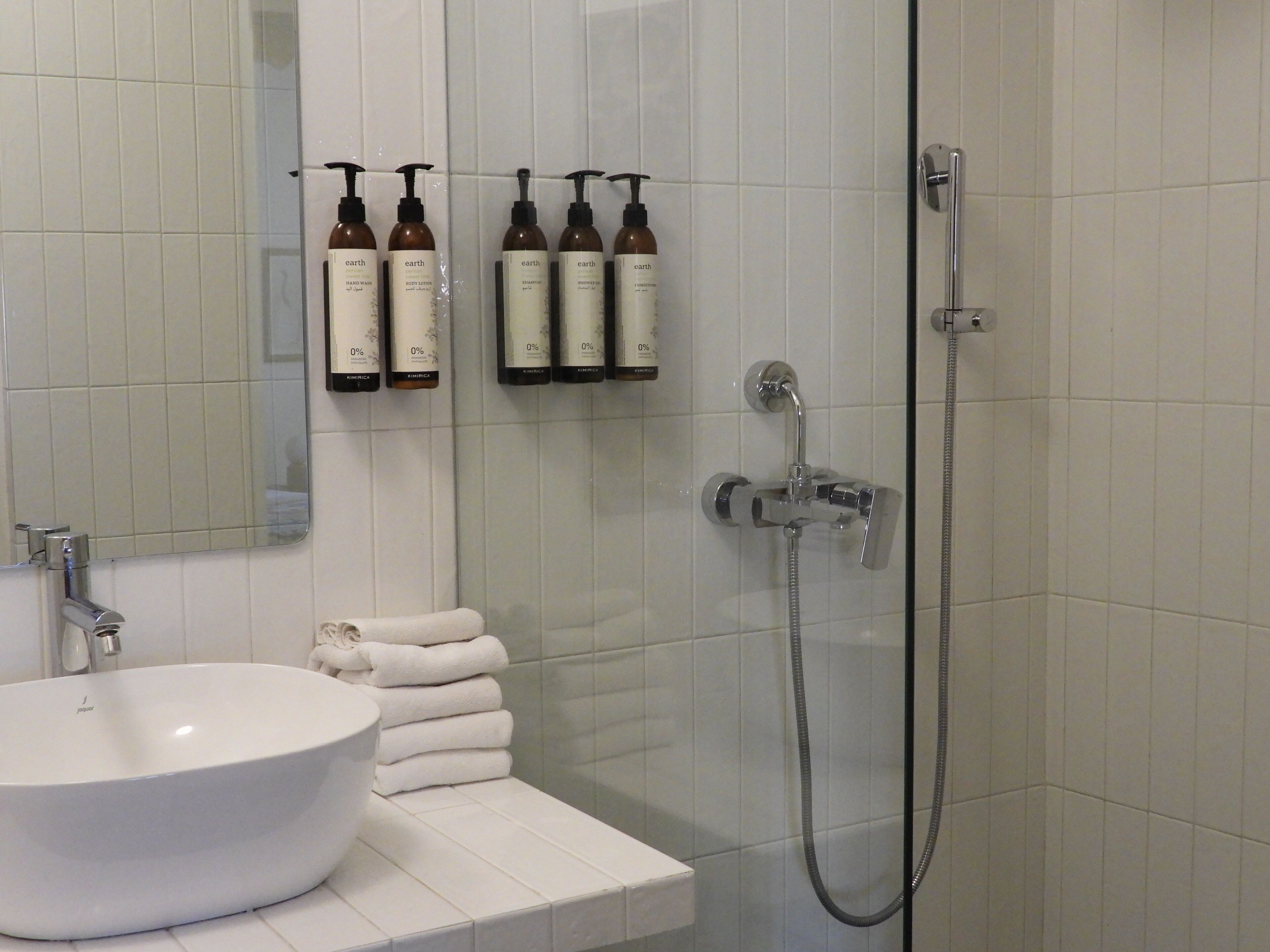 Un baño con un lavamanos blanco, un espejo y una ducha con cabezal de ducha manual. Hay botellas de champú, acondicionador y crema corporal en la pared.