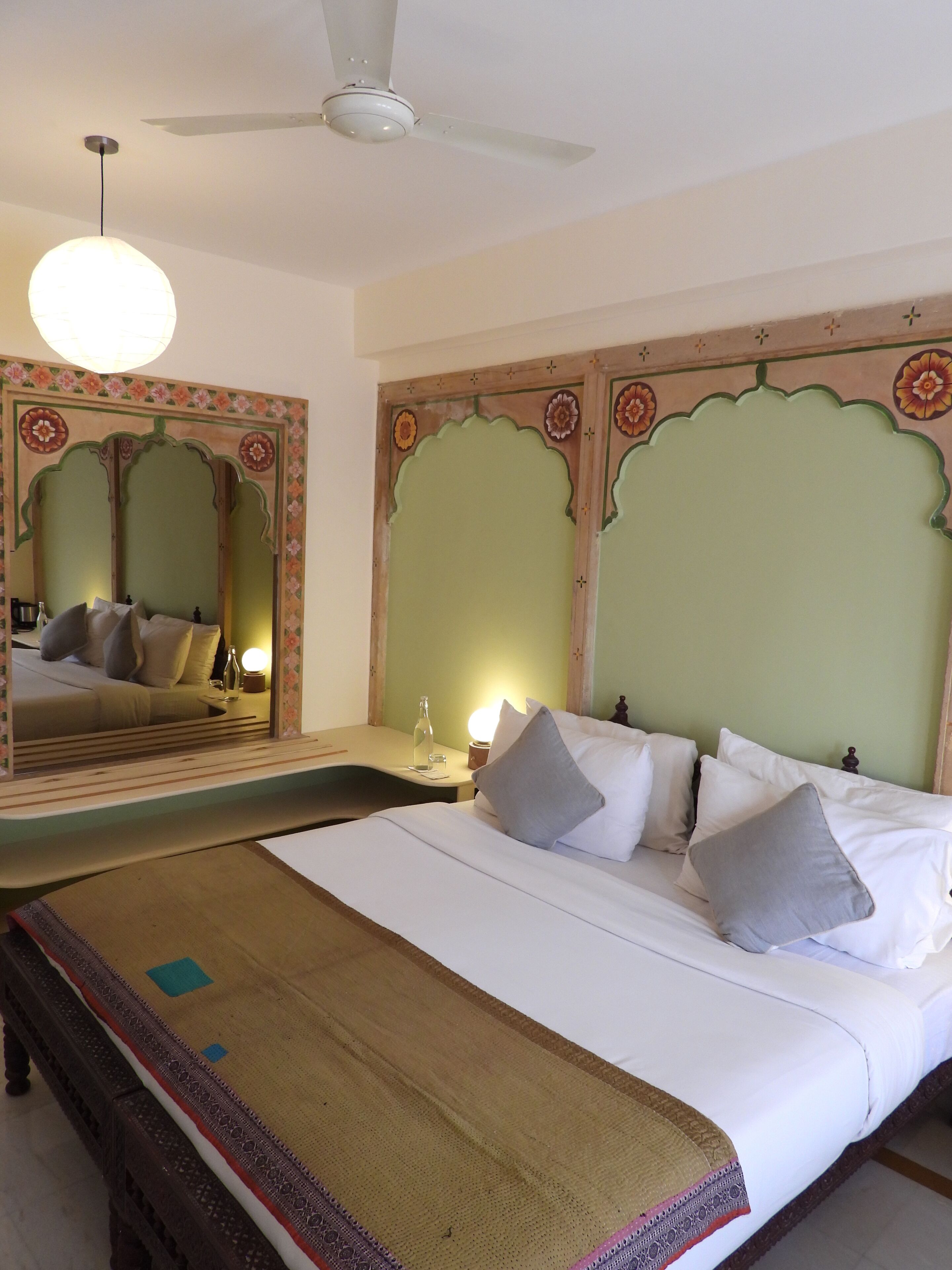 A hotel room with a bed, a nightstand, a mirror, a ceiling fan, and a decorative wall panel.