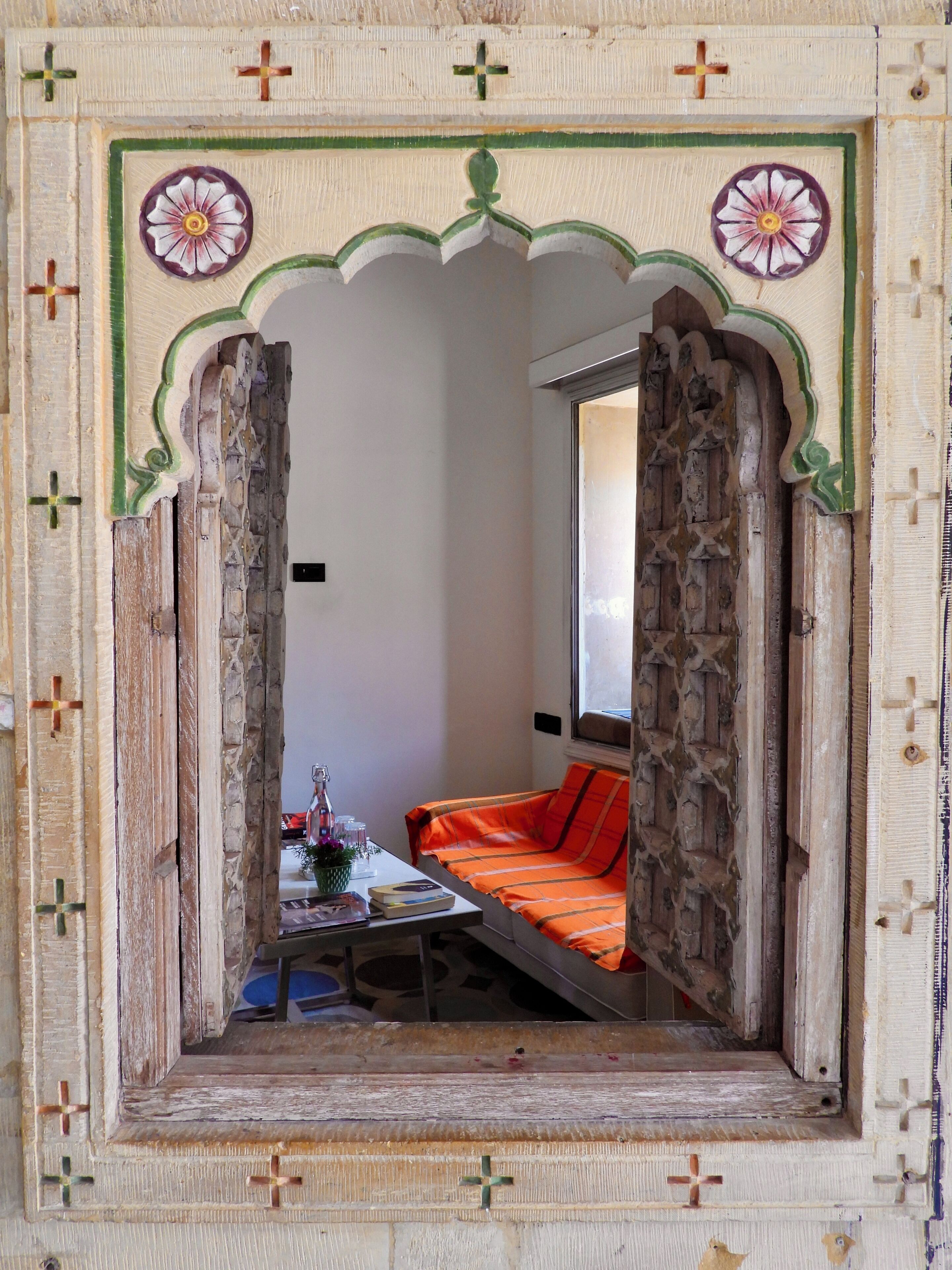 Een kamer met een traditionele boog die leidt naar een bed met oranje beddengoed, een klein tafeltje met een vaas en boeken, en een raam met uitzicht.