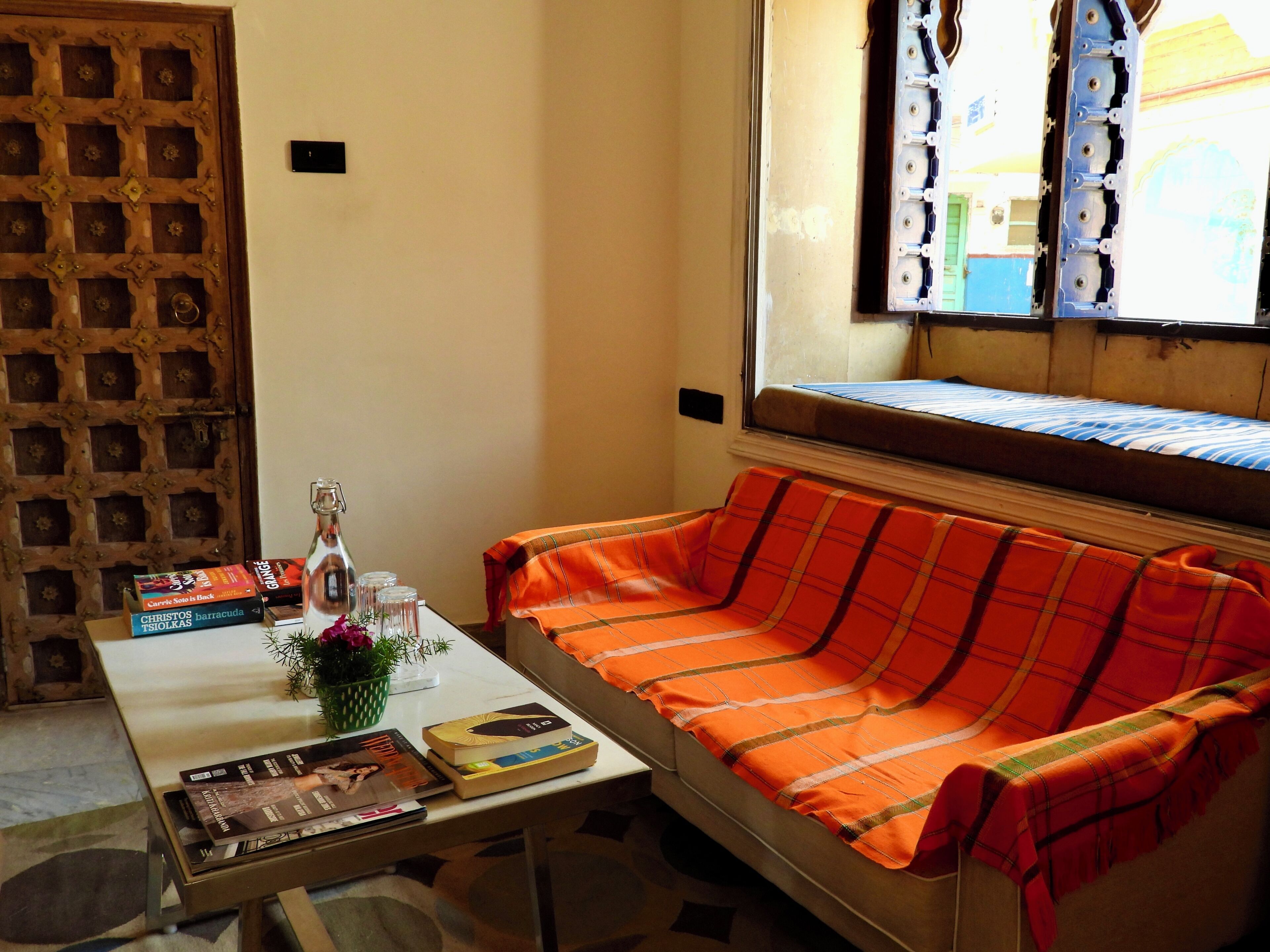 A room with a sofa, a table with books and a bottle, a window with blinds, and a wooden door.