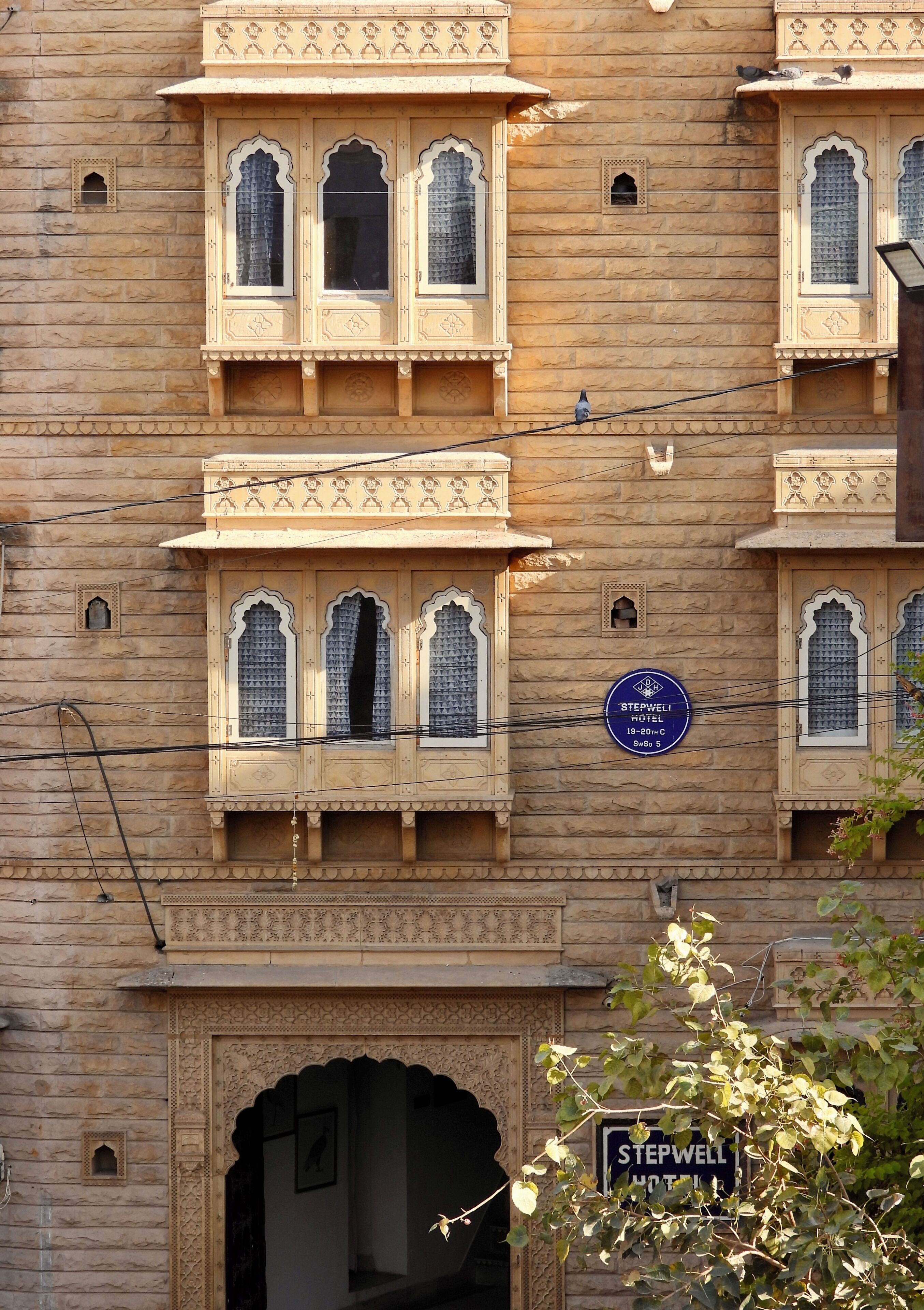 A building with traditional architecture, featuring arched windows and decorative elements. A signboard indicates the name "STEPWELL HOTEL" and a price range of 19-20C.