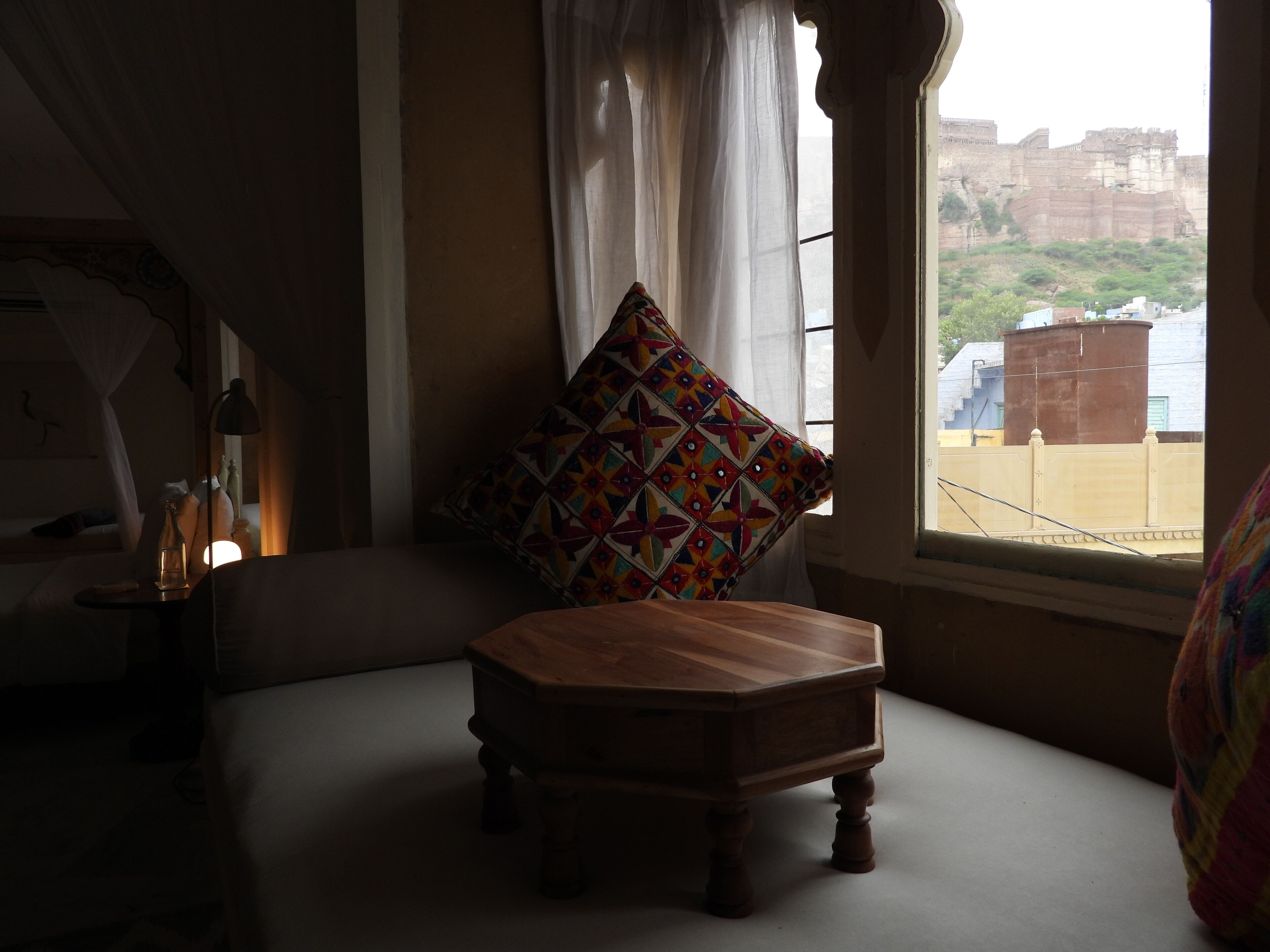Een kamer met een houten bankje, een kleurrijk kussen en uitzicht op een historisch fort door een raam.