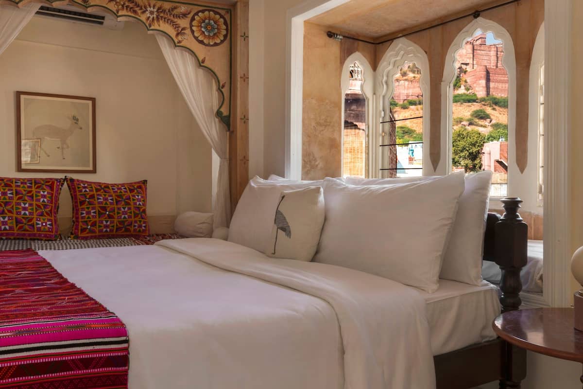 A bedroom with a bed, a bedside table, a patterned blanket, a framed picture, and a window with a view of buildings.