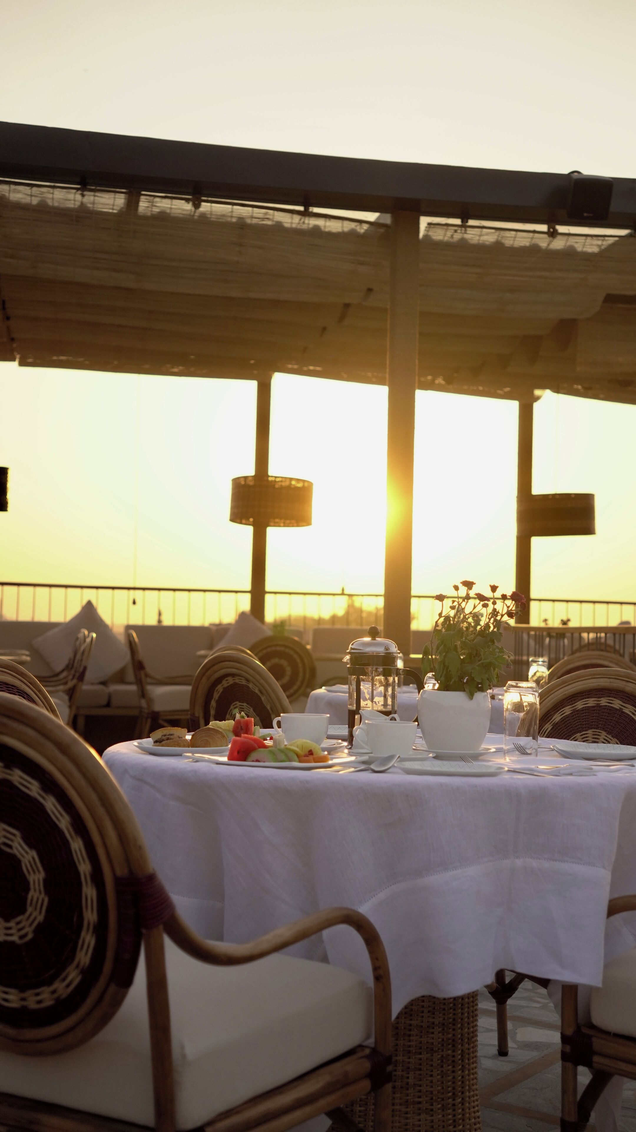 A table set for a meal with a view of the sunset, wicker chairs, and a cross-shaped structure in the background.