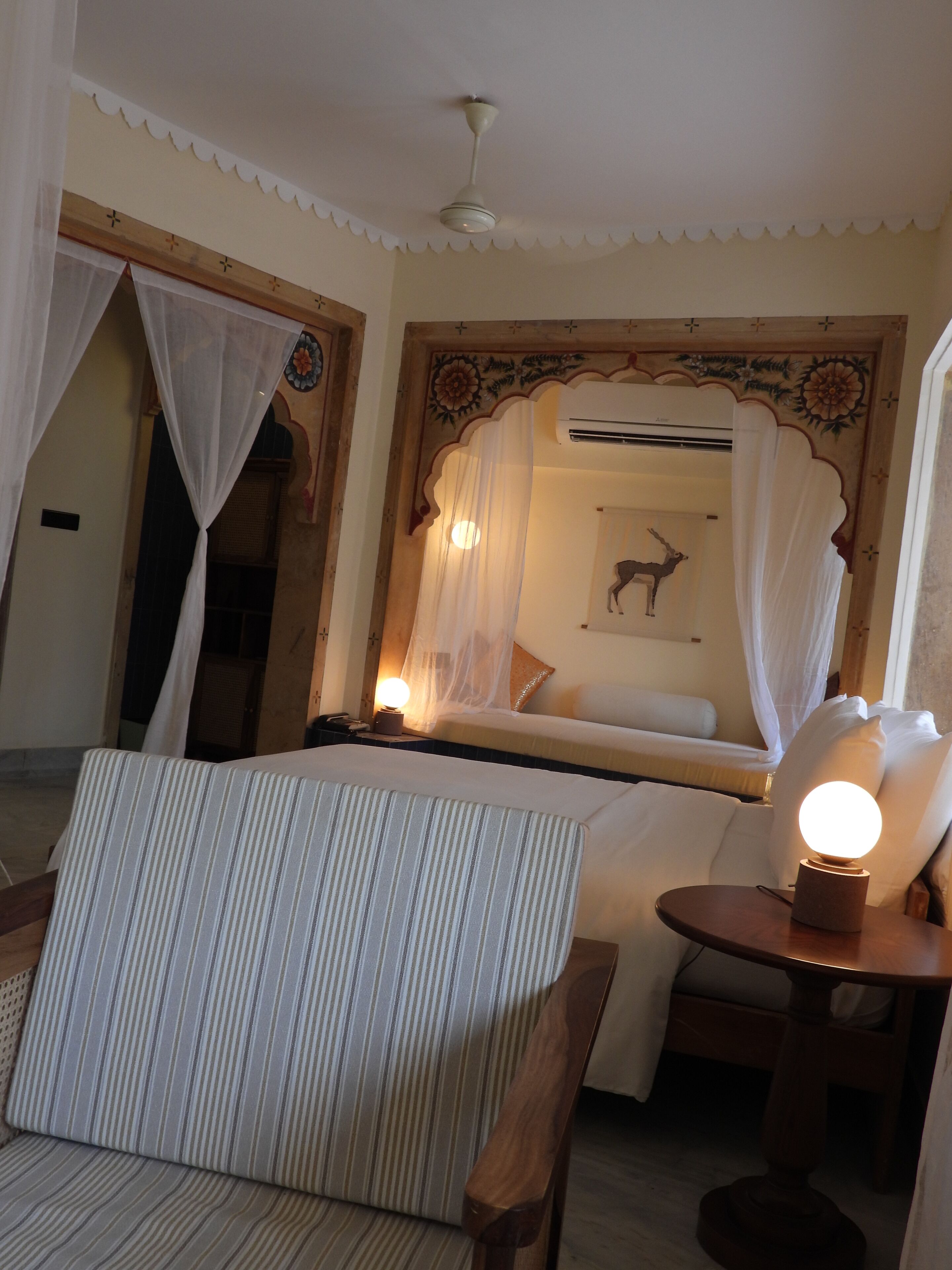 Une chambre d’hôtel comprenant un lit, une table de chevet avec une lampe, une chaise et un mur décoratif orné d’un motif floral.