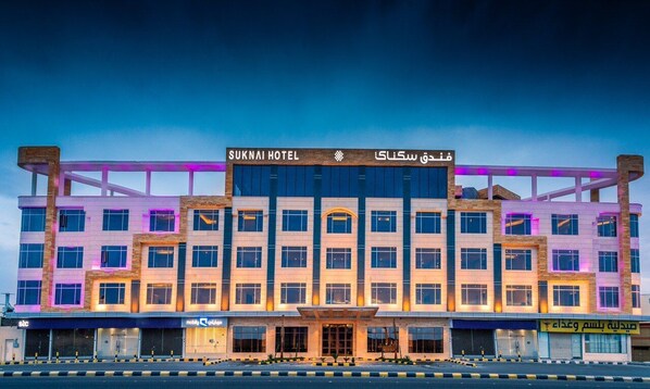 Un hotel de diversos pisos amb un cartell prominent que diu "SUKNAI HOTEL" i text àrab.