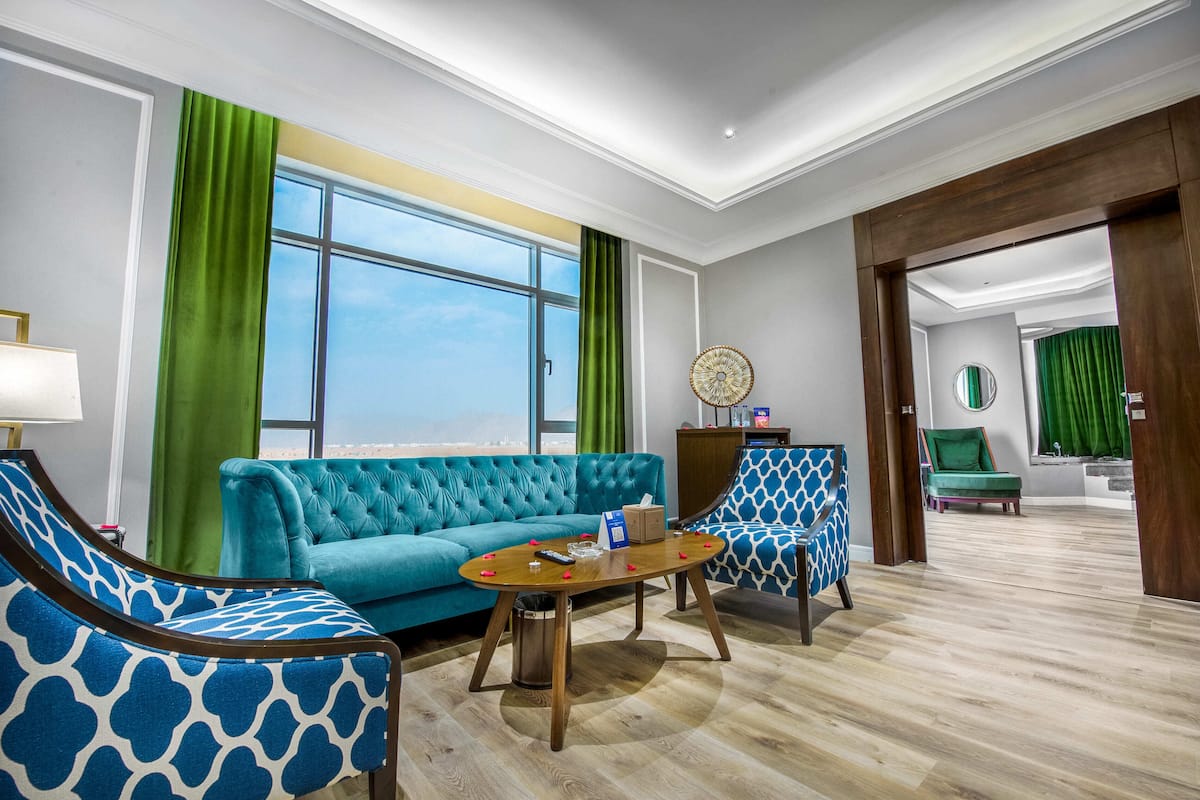 Una sala de estar moderna con un sofá verde azulado, sillones estampados, una mesa de centro de madera y vistas a la ciudad.