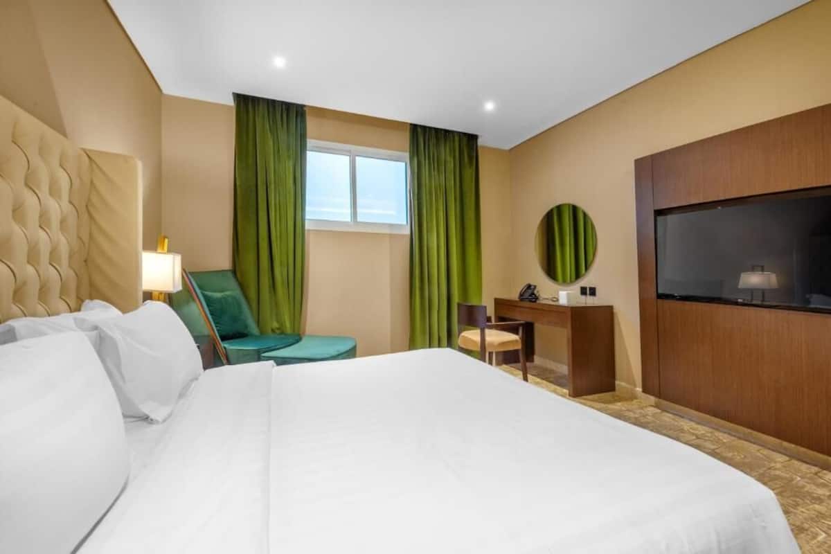 Una habitación de hotel con una cama grande, un escritorio, una silla, un televisor y una ventana con cortinas verdes.