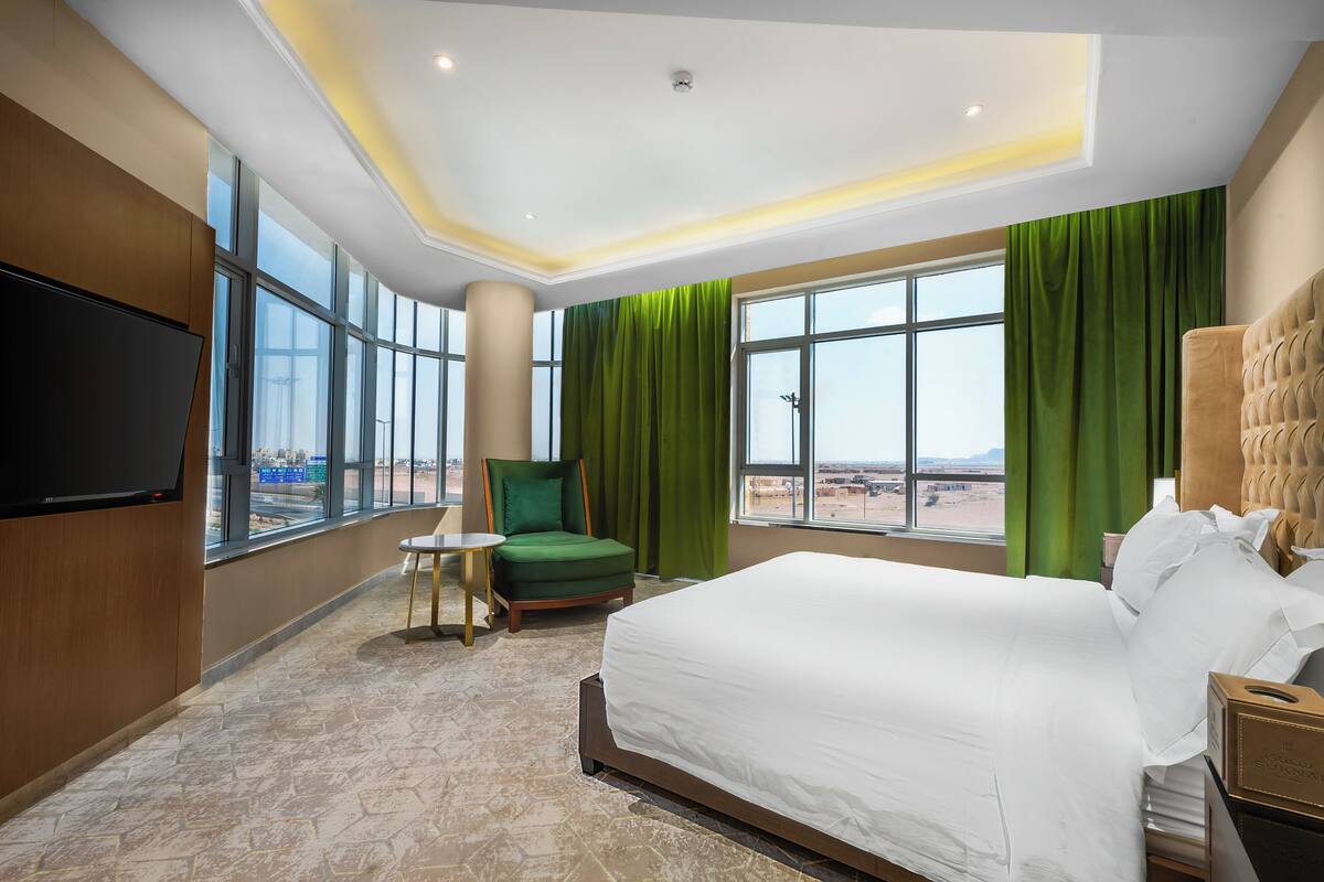 Habitación de hotel con una cama grande, un sillón verde, una mesita y un televisor.