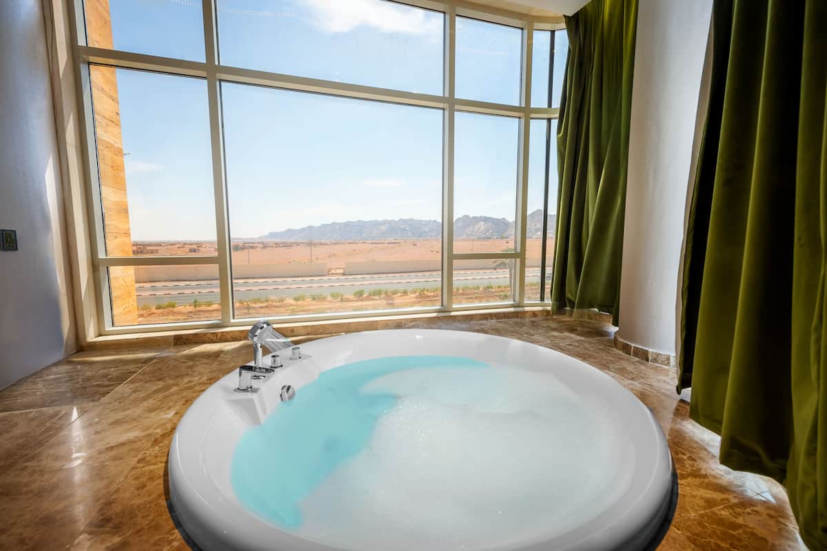 Un baño con una bañera grande, vista a un paisaje desértico a través de un ventanal amplio y cortinas verdes.