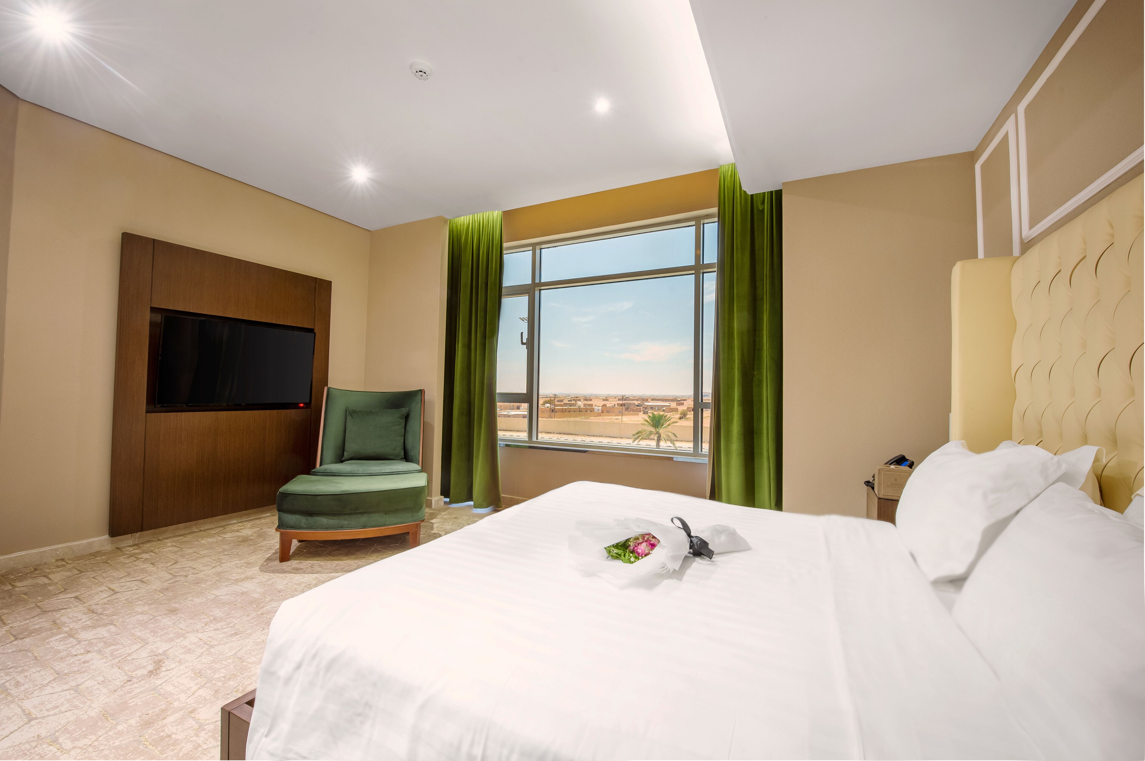 A hotel room with a bed, a green armchair, a TV mounted on the wall, and a window with curtains.